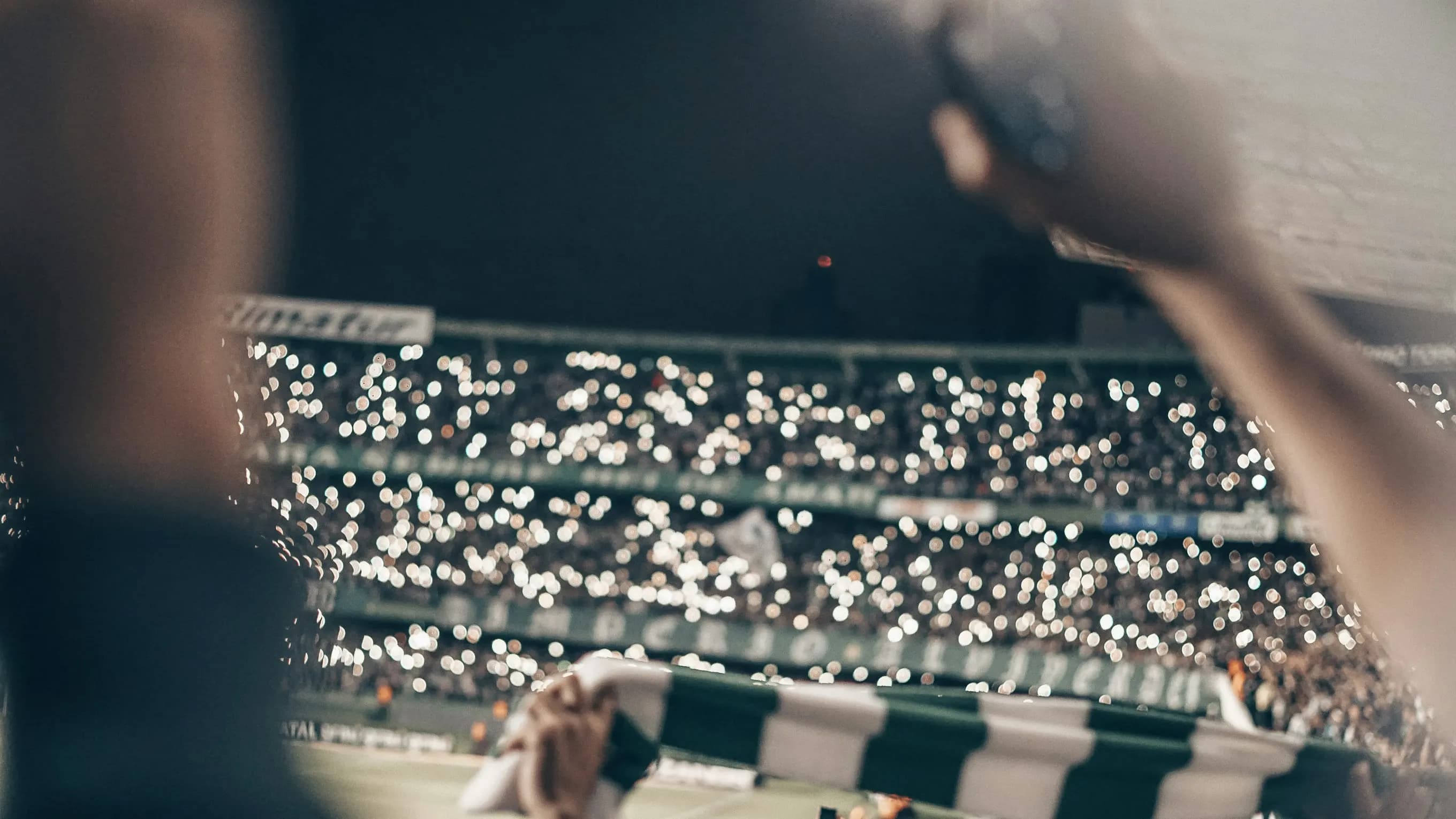 A football stadium at night lit by thousands of phone lights, a fan’s raised hand and scarf in the foreground